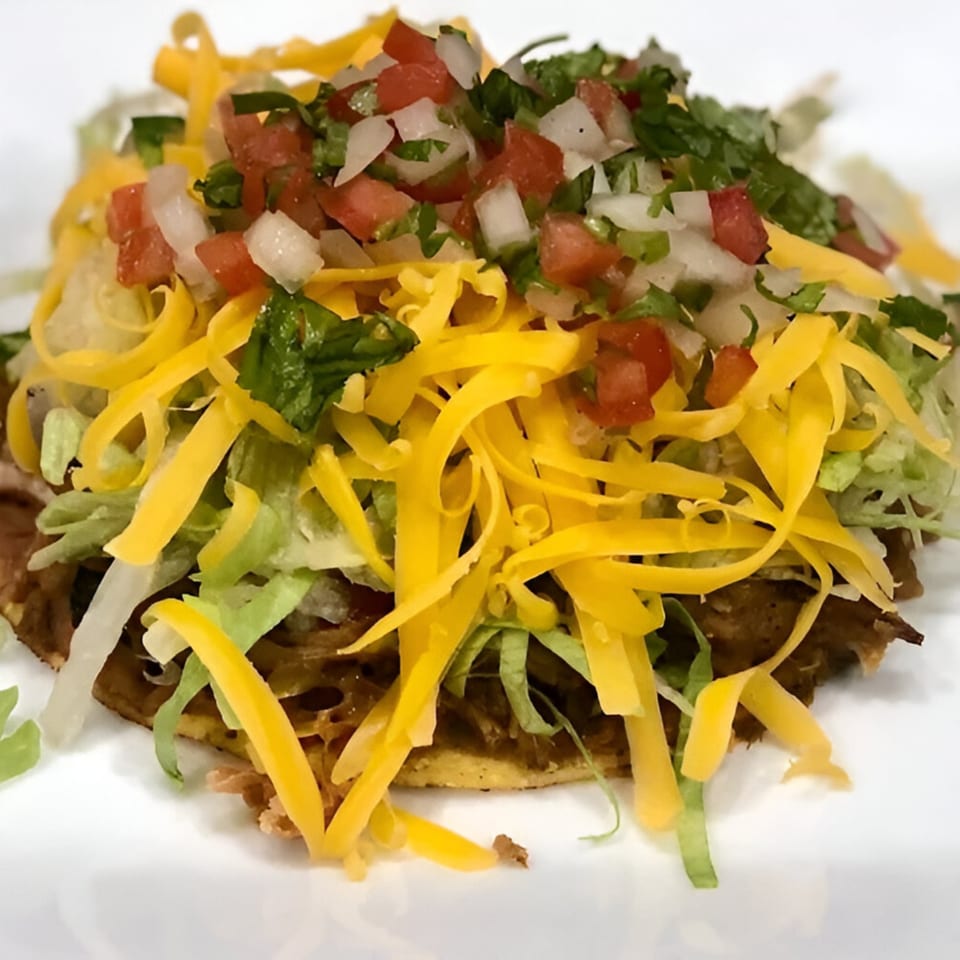 A Mexican tostada topped with shredded cheese, lettuce, and pico de gallo on a white plate.
